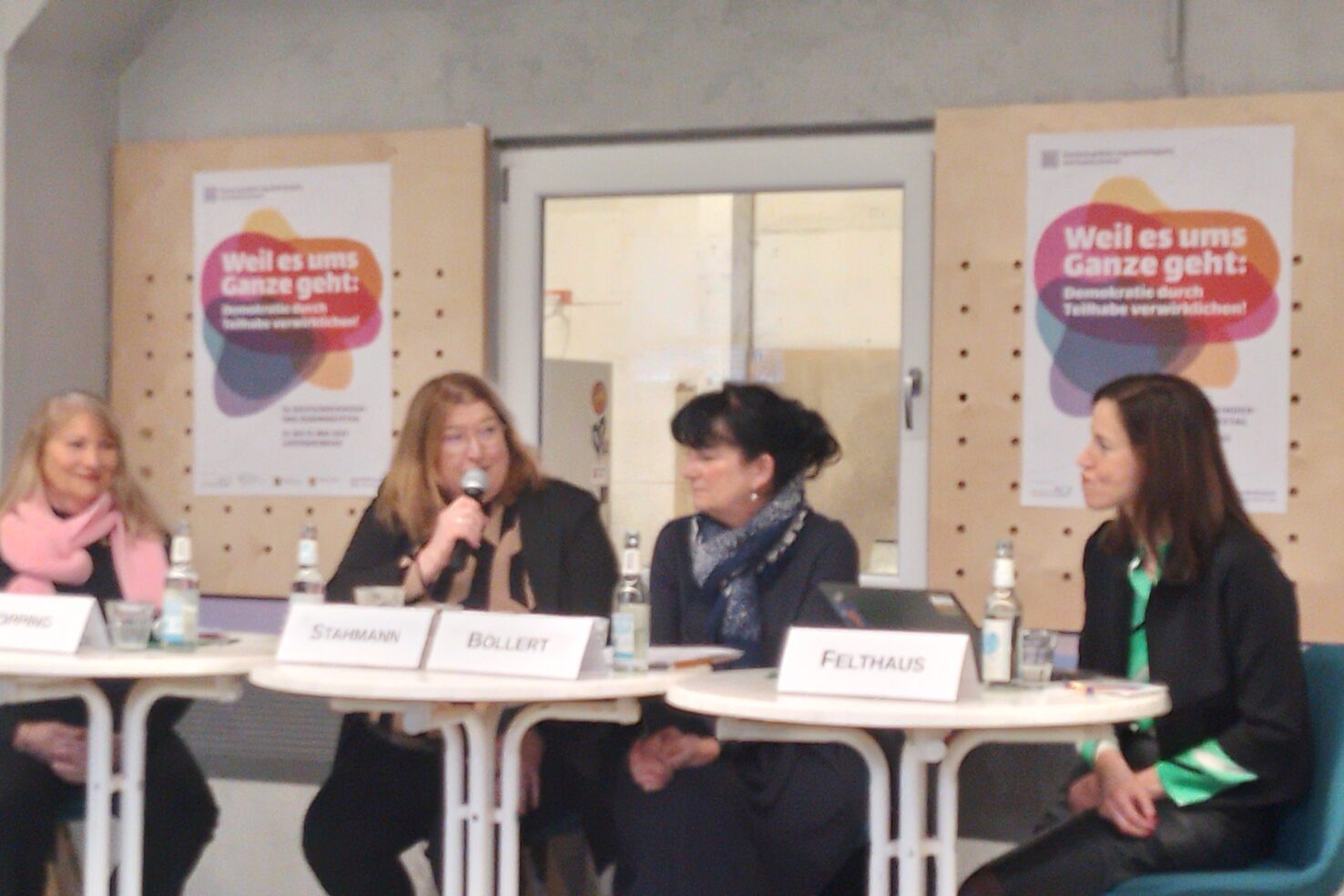 Vier Personen sitzen an Tischen und diskutieren. Vor ihnen stehen Namensschilder. Eine Person hält ein Mikrofon in der Hand.
