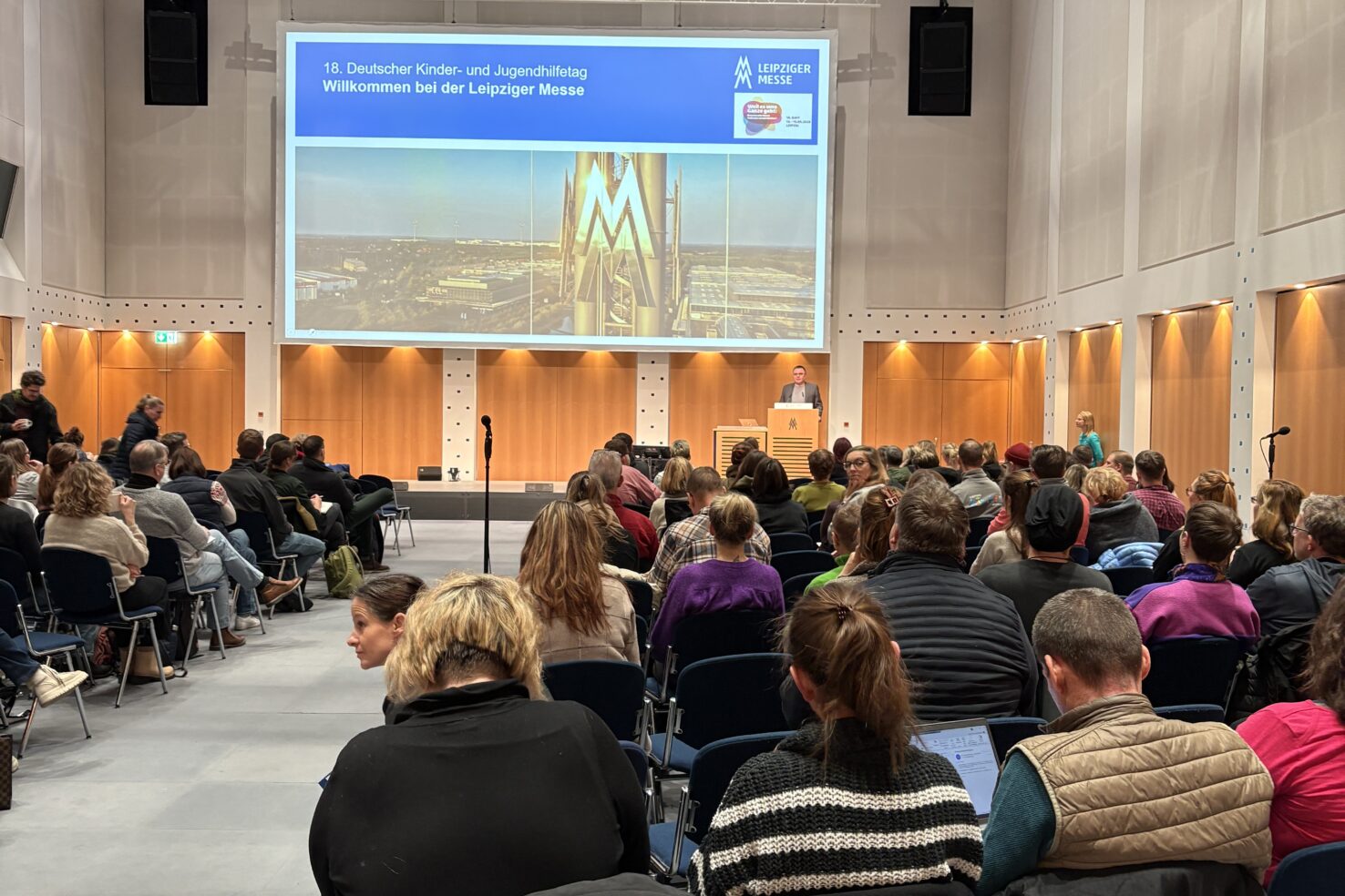 Ein Saal der Leipziger Messe ist gefüllt mit Menschen. Vorne steht ein Mann und hält einen Vortrag und hinter ihm läuft eine Power Point Präsentation.