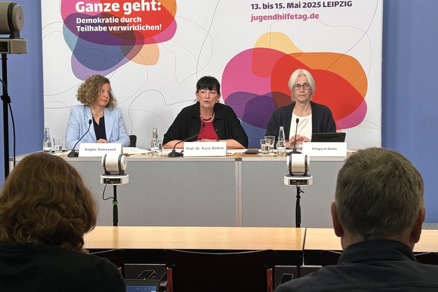 Drei Personen sitzen an einem Podium und haben Mikrofone vor sich. Im Hintergrund ist eine Pressewand des Kinder- und Jugendhilfetag zu sehen.