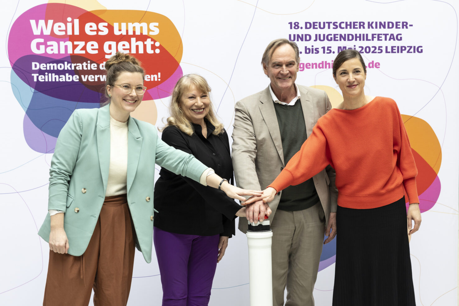 Vier Personen legen Hände übereinander auf einen roten Knopf. Im Hintergrund sind Schriftzüge des deutschen Kinder- und Jugendhilfetag zu sehen.